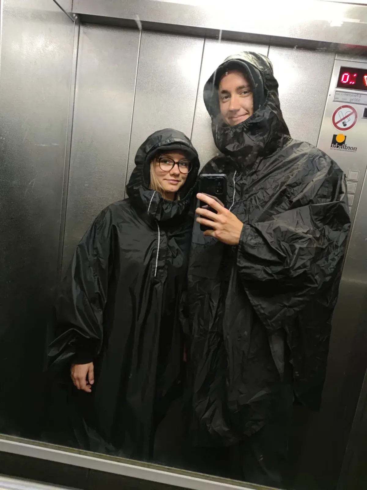 Erik and Darja in black rain ponchos, a mirror selfie in a lift